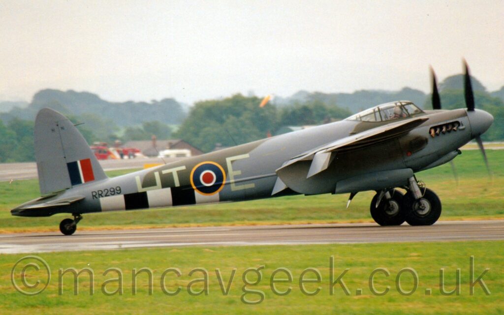 Side view of a twin propellor-engined, World War 2-era bomber aircraft taxiing along a runway from left to right. The plane is covered in an olive-green and grey camouflage scheme, with thick black and white bands around the lower rear fuselage. There are very pale letters "HT-E" either side of a blue, white, and red roundel outlined in yellow. There is a blue, white, and red fin flash on the lower part of the tail, above the black serial "RR299" on the upper rear fuselage. Lush green grass fills the foreground, with more grass leading up to a house and a fire truck in the distance, with trees beyond, under hazy grey skies.