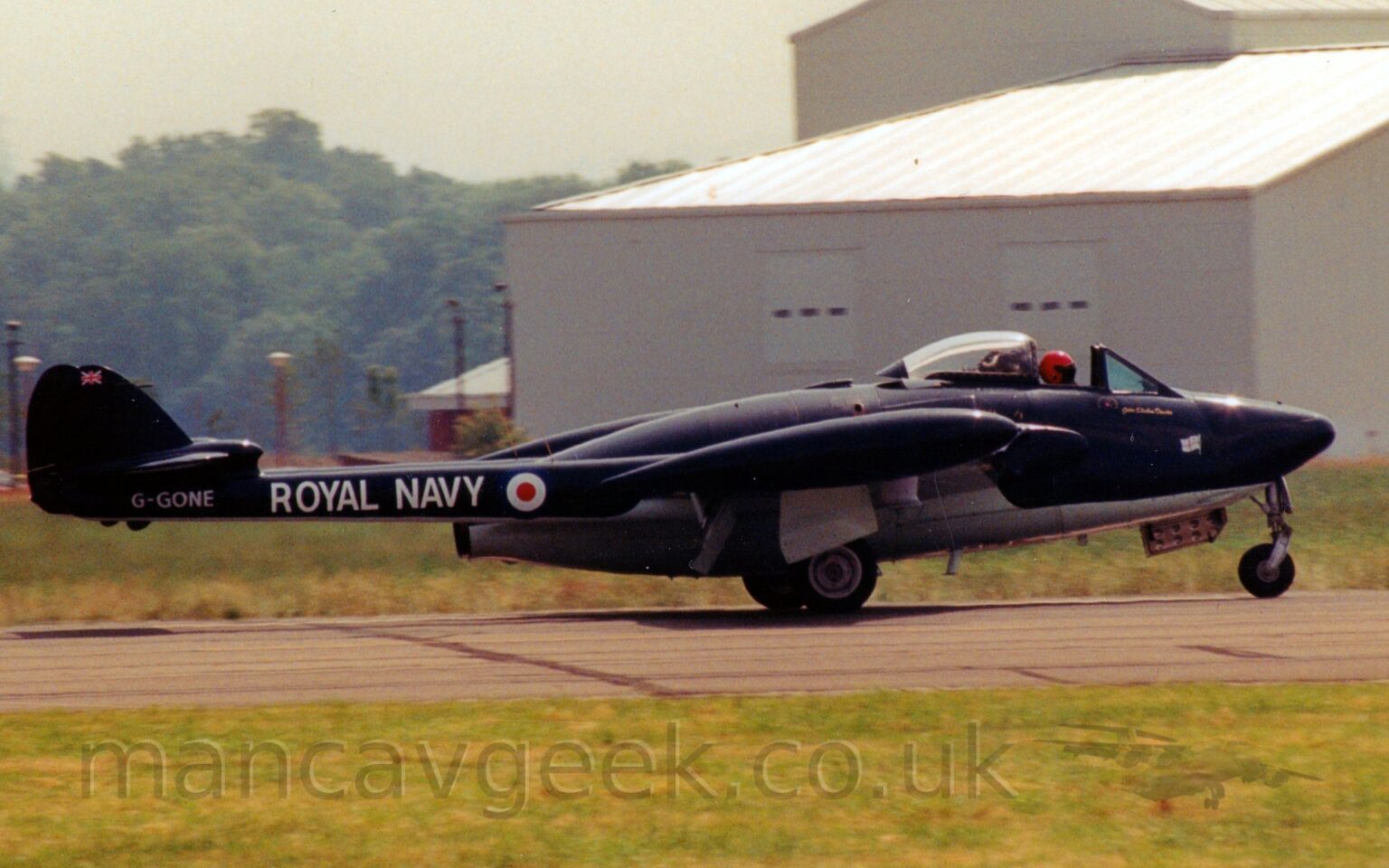 Side view of a black, single engined military jet fighter taxiing from left to right along a grey runway. The plane is basically a large black engine pod with a white belly, with twin black tail booms stretching backwards from the wing root, carrying the small twin tails. The cockpit canopy at the front of the plane is open, slid back along the top of the fuselage, with the bright red helmet of the pilot clearly visible in the gap. The tail booms has white "Royal Navy" titles, just aft of a blue, white, and red roundel. The registration "G-GONE" is on the end of the boom in white, under the tail. Green grass fills the foreground, with a large grey building dominating the frame on the right, and trees on the left in the background, under bright but hazy skies.