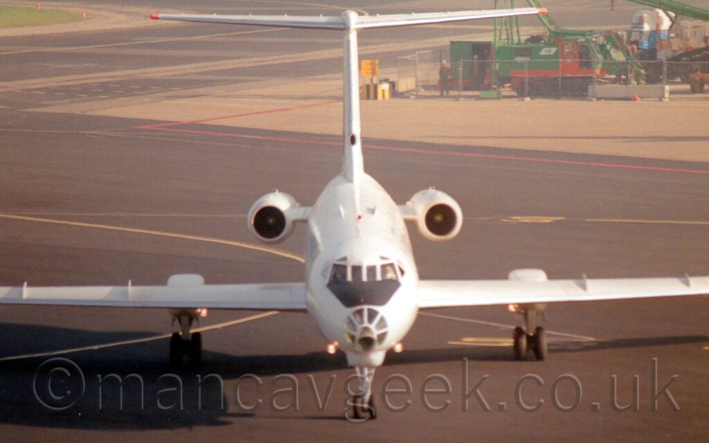 Head-on view from slightly above of a twin engined jet airliner with the engines mounted on the sides of the rear fuselage, taxiing pretty much straight towards the camera. The plane is almost entirely white, with only a black anti-dazzle panel on in front of the cockpit, above the heavily glazed nose-cone. The main landing gear is obviously in a strange place, attached to (and retracting into) pods mounted partway along, and at the rear of, the wings, rather than retracting into the fuselage as on most airliners. Black tarmac apron marked with yellow and red lines fills most of the background, with a building site on a grey concrete apron in the top right corner.