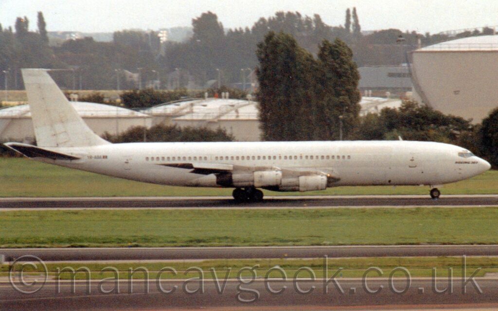 Side view of a s4-engined jet airliner moving from left to right along a black runway, with flaps deployed from the rear of the wing, and more drooping down over the front, suggesting this plane has just landed. The plane is almost entirely white, with only black text "Boeing 707" on the nose, and the black registration "YR-ABA" next to a small Romanian flag adding any contrast. Black strips of taxiway alternate with wider strips of green grass in the foreground, with more grass and trees in the background leading up to squat, white, domed fuel tanks in the distance, with more trees vanishing into haze beyond that.