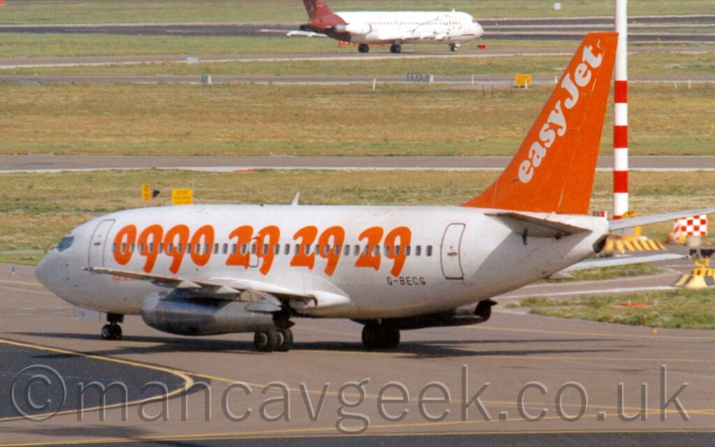 Side view of a twin engined jet airliner taxiing from right to left along a grey taxiway. The plane is largely white, with a large, orange telephone number emblazoned on the side of the body, covering the passenger cabin windows, with the black registration "G-0BECG" on the lower rear fuselage. The tail is bright orange, with white diagonal "EasyJet" titles along the leading edge. Large areas of green grass fill the background, crisscrossed with grey taxiways, and a white and mauve jet airliner taxiing to the right at the top of the frame.