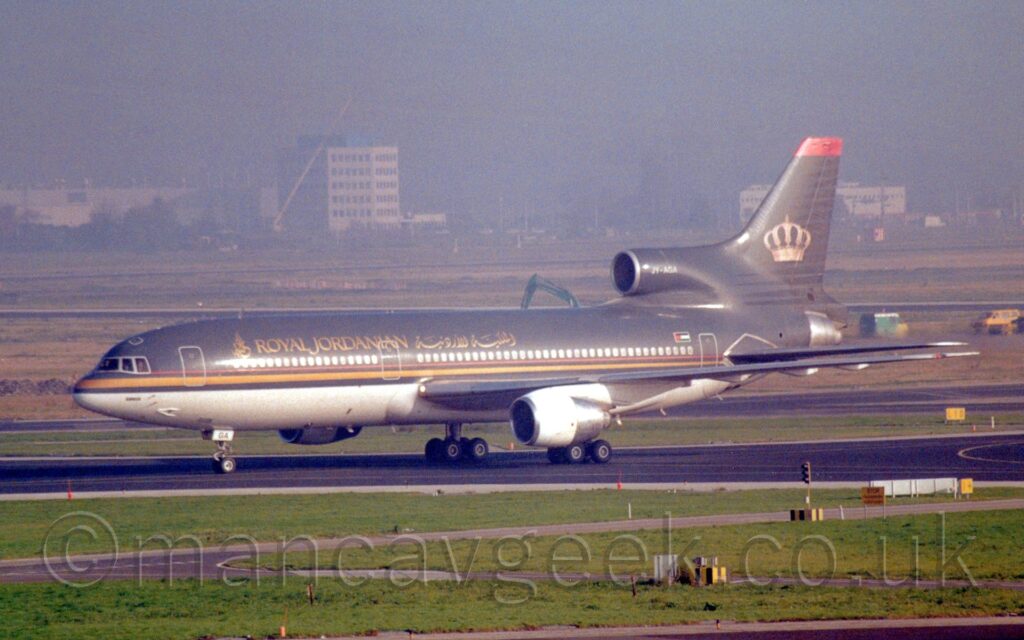 Side view of a 3 engined jet airliner taxiing from right to left along a black taxiway. The plane is mostly a deep purple, verging on black, with a thin red and thick gold stripe running along the body, and a light grey belly and engines. There are gold "Royal Jordanian" titles on the upper forward fuselage, with golden Arabic titles aft of that. There is a small Jordanian flag on the upper rear fuselage, , and the grey registration "JY-AGA" on the side of the centre engine intake in front of the tail. The tail itself has a large golden crown, and a red stripe right at the top. Green grass criss-crossed with grey roadways fill the foreground, with more grass leading up to taxiways and a runway in the background, and tall buildings vanishing in to haze in the distance.