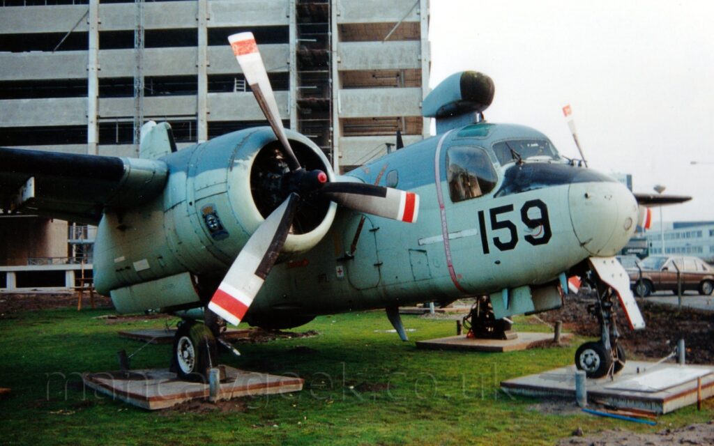 Front side view of a high-winged, twin propellor-engined military aircraft standing on 4 wooden platforms in the middle of a grassy area, on display outside a museum. The plane is mostly a very pale creamy blue, with dark grey upper surfaces, the large black number "159" on the nose under the cockpit, and a red band around the fuselage, level with the propellors, immediately aft of the cockpit. There is a small, grey radome with a black front piece on top of the fuselage, between the cockpit and wing. A tall grey building is under construction in the background on the left, with a brown car and a lower grey building on the right.