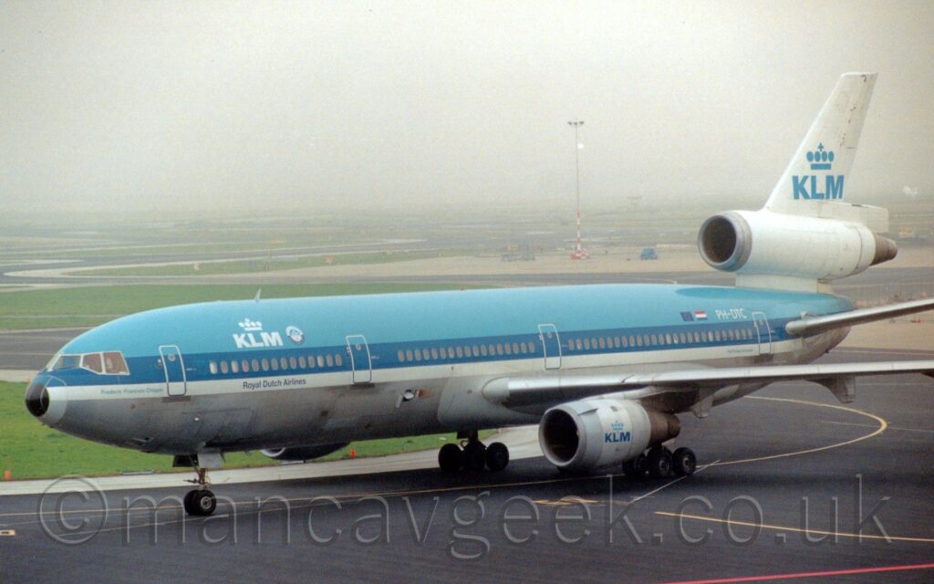 Side view of a 3-engined jet airliner taxiing from right to left, following a yellow line marked on a black taxiway. The plane is blue un top, with a grey lower half, and a thick dark blue stripe running along the body, covering the passenger cabin windows, with a thinner white stripe just below that. There are white "KLM" titles on the upper forward fuselage, under a stylised white crown, with small blue text "Royal Dutch Airlines" on the white stripe below. The white registration "PH-DTC" is on the upper rear fuselage, just aft of Dutch and EU flags. The tail is white, and has the centre engine running straight through the bottom, with blue "KLM" titles under a stylised blue crown on the top section. This logo is repeated on the grey engine pods mounted under the wings, although this time with the "KLM" text in a darker blue. Black taxiway fills the foreground, with islands of green grass surrounded by taxiways meandering through the background, slowly being eaten up by mist in the distance.