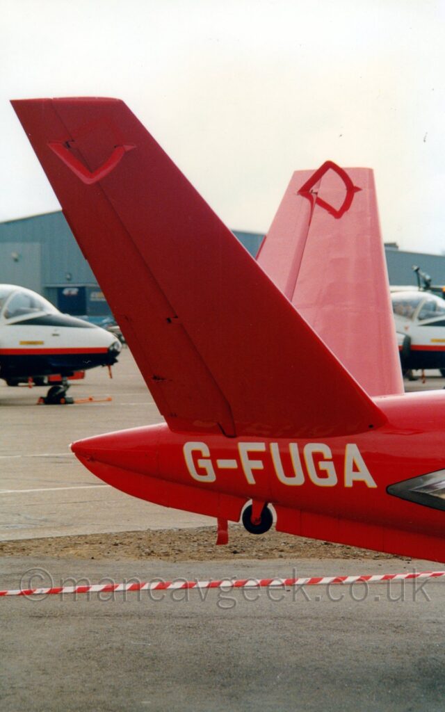 Vertically oriented close-up of the twin red V-Tails on the end of the fuselage of a red aircraft. The white registration "G-FUGA", is under the tail above a circular rubber bumper. In the foreground, red and white striped tape stretches across the frame, separating the camera from the plane, above grey tarmac. The cockpits of a pair of white and dark blue planes can be seen in the background, with a large grey hangar beyond that, under bright but hazy sky.