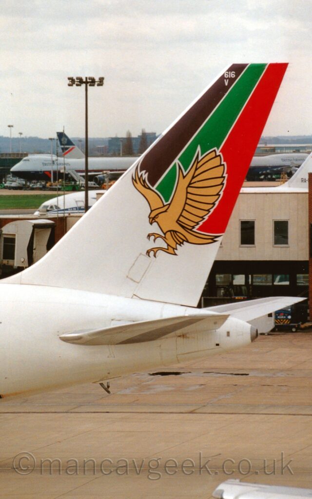 Vertically oriented close-up of the tail of a jet airliner taxiing from right to left. The plane is mostly white, an image of a golden falcon with wings raised and legs extended as if to attack, on the tail. Behind this bird, diagonal brown, green, and red stripes fill the top of the tail, with the white number "616" right at the top, above the white letter "V". Grey concrete image fills the bottom of the image in the background, with a creamy-brown terminal building stretching across the frame from the right, and a white jet airliner partially hidden behind that. 4 very large, 4-engined jet airliners in a grey and dark blue livery are in the distance, trying to hide behind the tail of this plane, with buildings beyond them, under cloudy grey sky.