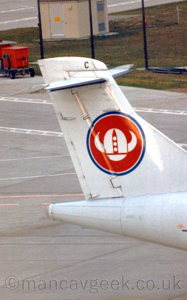 Vertically-oriented close-up of the tail of a white airliner taxiing from left to right. The white tail has a red circle outlined in blue, with a stylised white Viking helmet in the middle, and the black letter "C" right at the top of the tail. Grey concrete apron marked with white lines fills the background, with a grassed area behind a black mesh fence in the top right corner, and a red box on wheels in the top left corner.