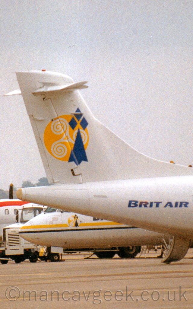 Vertically oriented close-up of the rear fuselage and tail of an airliner parked facing to the right. The plane is mostly white, with a yellow circle on the tail, with a small white triangle in the middle and a white spiral coming off each corner, running counter-clockwise. Next to this is a large blue arrowhead pointed upwards, with 3 small blue diamonds around the top. Grey concrete apron fills the bottom of the screen, with the nose and lower fuselage of another airliner visible under the tail of this plane in the background, a small white tug beyond that, and a 3rd white airliner on the left of the frame behind that. Flat grey sky fills the rest of the frame.