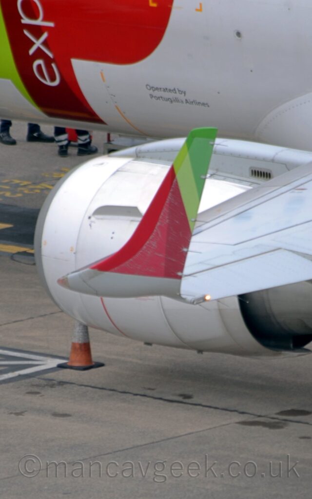 Close-up of the left upturned wingtip of a white, twin engined jet airliner parked facing to the left. The wingtip has red, light green, and dark green stripes, and is extremely faded towards the rear edge. The engine pods are white, and slung low under the wings. There is a light green and red logo on the plane's forward fuselage, with the white letters "exp" overlaid vertically. There are black "Operated by Portugalia Airlines" titles just forward of the wing root. Grey concrete apron fills the rest of the bottom of the frame.