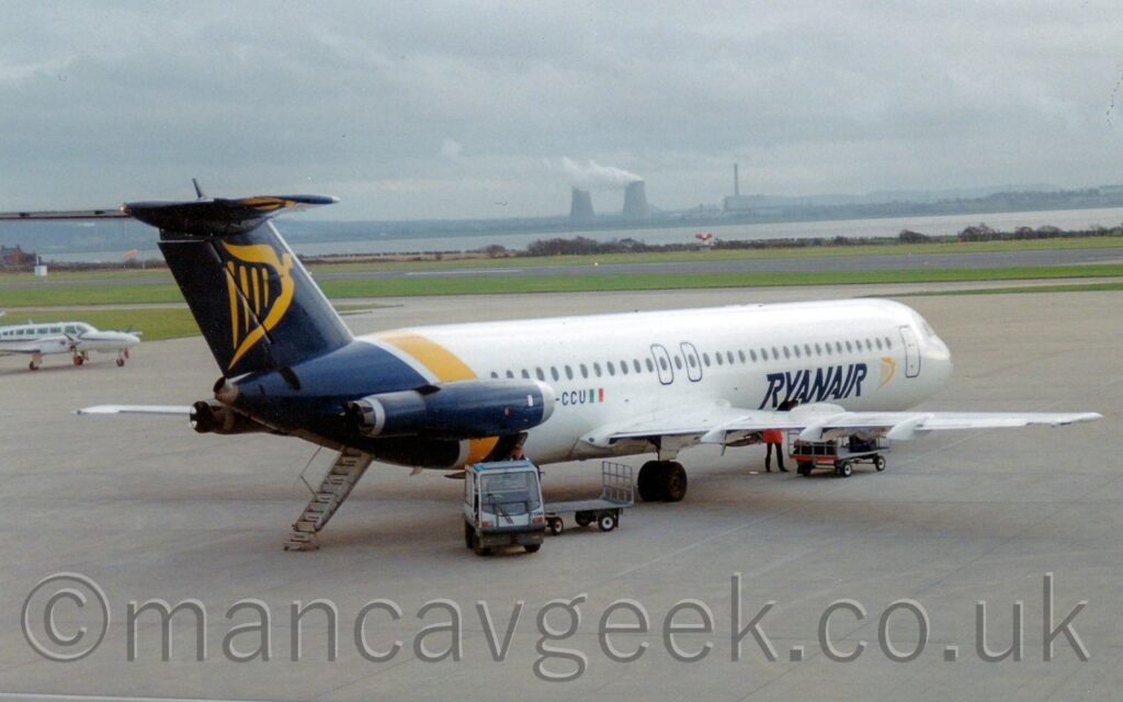 Side view of a white, twin engined jet airliner with the engines mounted on the sides of the rear fuselage, parked facing to the right and slightly away from the camera. There are dark blue "RyanAir" titles on the lower forward fuselage next to a yellow, winged Irish harp. A larger version of the harp is on the dark blue tail/ The rear fuselage is the same dark blue, with a diagonal yellow stripe in front, with the dark blue engine pods mounted on the sides. The registration "EI-CCU" is on the side of the rear fuselage, partially obscured by the engine pod. A luggage cart with a couple of trailers is parked in front of the plane, with only a couple of cases on the rear trolley. Grey concrete apron fills most of the frame, with a white twin engined BizProp parked on the left edge of the frame. Grass in the background leads up to a wide river in the distance, with an industrial landscape on the far side, under grey sky.