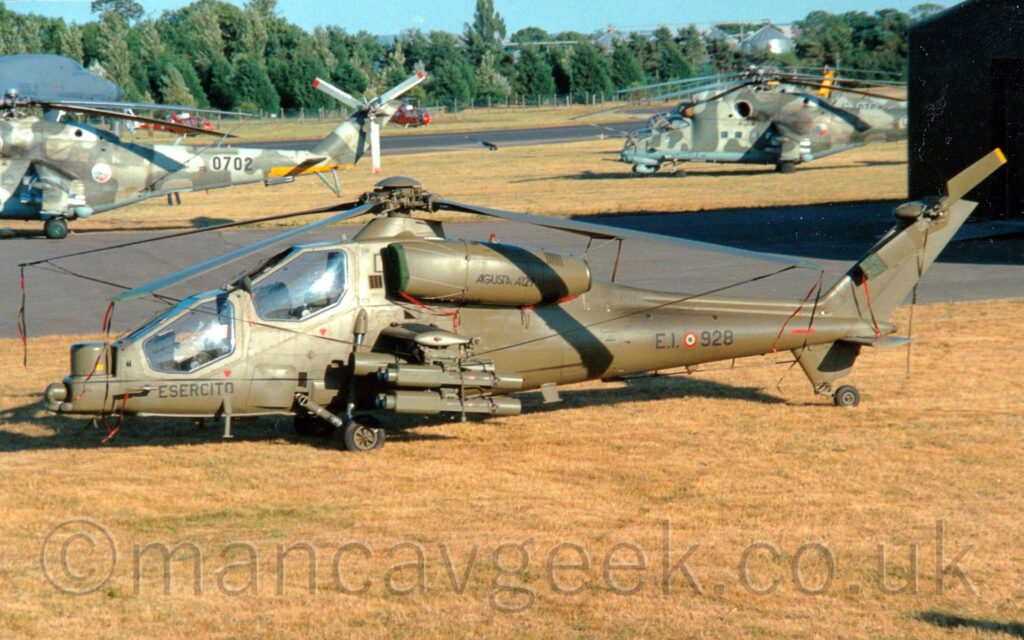 Elevated side view of a military attack helicopter with 2 staggered cockpits, one above and behind the other, in an olive-green paint job, parked on grass facing to the right. There are black "Esercito" titles under the forward cockpit, "EI" and "928" either side of a red, white, and green roundel on the rear fuselage, and "AgustaA129" titles on the sides of the engine pods mounted on the sides of the upper fuselage, under the rotors. Stub wings below the engine pods hold 4 rocket pods. Yellowing grass fills the foreground. With black taxiways and more grass leading off to trees in the distance. A pair of larger attack helicopters in a stone-grey camouflage scheme are behind this helicopter, with a pair of smaller, red and white helicopters further back. Pale blue sky fills the top of the image.
