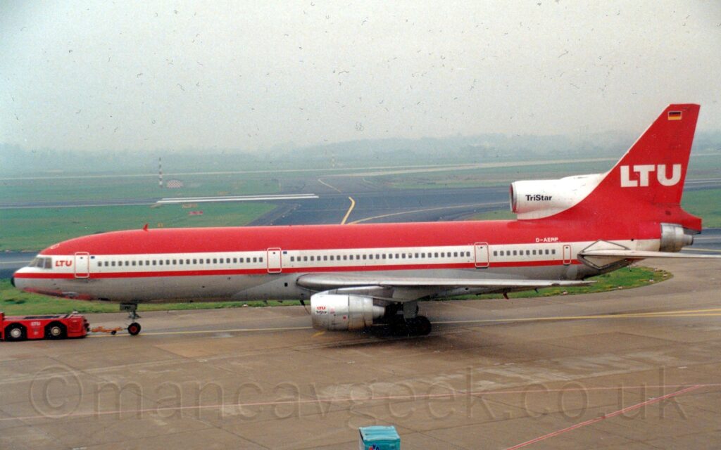 Side view of a red and white, 3 engined jet airliner being towed from right to left by a low-slung, red tug attached to the nose-wheel by a along yellow pole. The plane has red upper surfaces with a metallic grey belly, with a thick white stripe running along the body covering the cockpit and passenger cabin windows. There are small, red, "LTU" titles on the upper forward fuselage, between the rear of the cockpit and the forward door. The red tail has large white "LTU" titles in the middle, with a small German flag at the top. The under-wing engine pods are a bare metal, with small, red "LTU" titles and black "TriStar" text next to the intake. The centre engine intake in front of the tail is white, with black "TriStar" text, Grey concrete apron fills the foreground, with green grass and black taxiways and a runway vanishing into mist in the background.