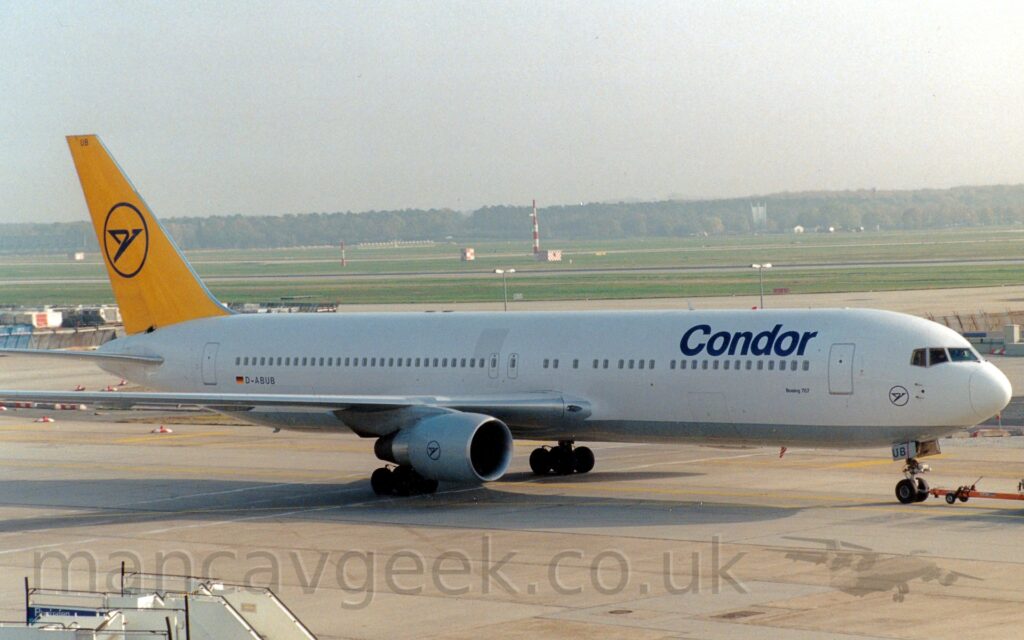Side view of a twin engined jet airliner bring towed from left to right by an out-of-frame tug attached to the nosewheel by a long orange pole. The plane is largely white, with dark blue "Condor" titles on the upper forward fuselage, and a grey belly, as well as the black registration on the lower rear fuselage, next to a small German flag.. The black outline of a circle containing a stylised dark blue flying bird is on the lower forward fuselage, under the rear of the cockpit windows, as well as on the grey engine pods under the wings. The same logo appears in a larger form on the bright yellow tail. Grey concrete apron fills the foreground, with a large grassed area crossed by A pair of dark grey runways, leading up to trees in the distance, under bright but hazy skies.