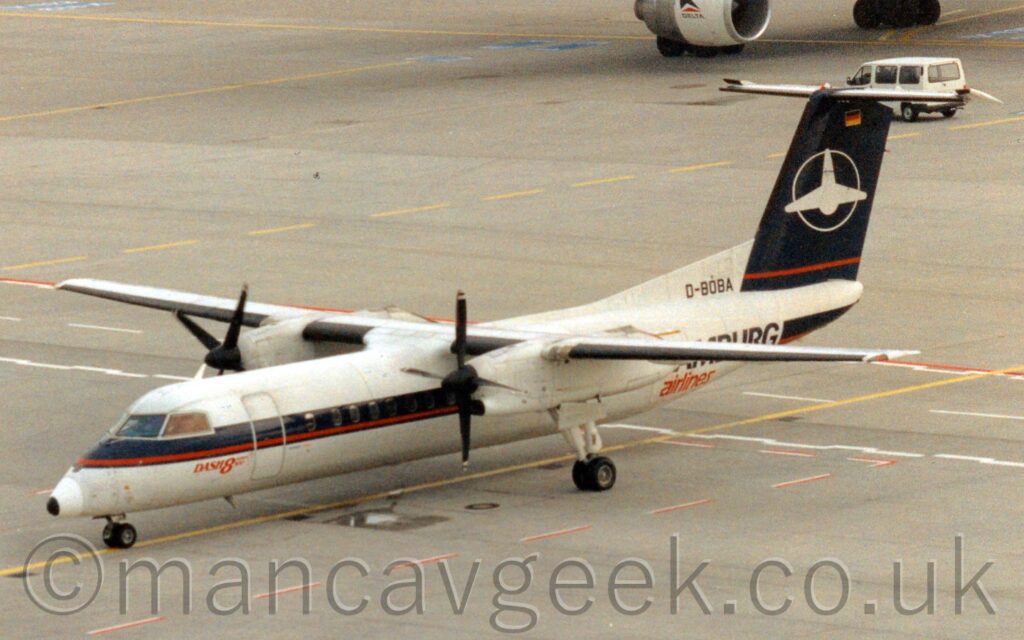 Elevated side view of a high-winged, twin propellor-engined airliner taxiing from right to left, following a yellow line marked on the grey concrete apron. The plane is mostly white, with a thick dark blue and thin red stripe running along the body, covering the passenger cabin windows. There are red "Dash8" titles in front of the forward passenger door, just below the rear of the cockpit windows. Black and red "Hamburg Airlines" are on the side of the rear fuselage, the first word partially obscured by the planes wing. The black registration "D-BOBA" is on a white fillet at the front of the tail. The tail is dark blue, with thin white outline of a circle around a highly stylised white aircraft design, a thin red stripe at the base of the tail, and a small German flag at the top. Grey concrete apron fills the rest of the frame, with the wheels and engines of another jet airliner just coming in to view in the top right corner.