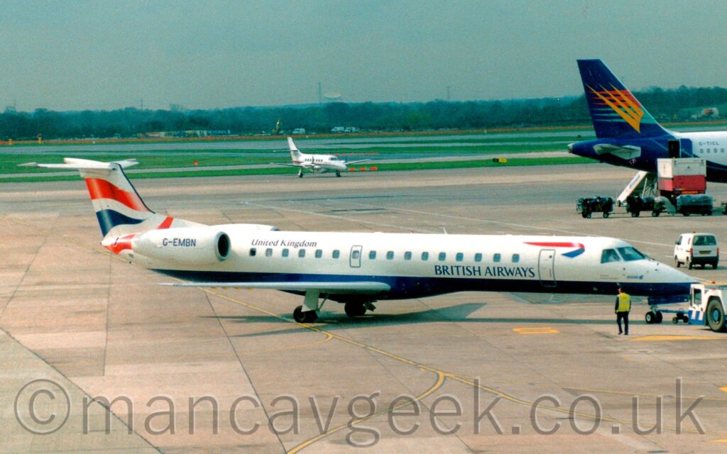 Side view of a white and blue, twin engined jet airliner with the engines mounted on the sides of the rear fuselage, facing to the right but being pushed back to the left by a low-slung white and blue tug attached to the nose-wheel by a long blue pole. The plane is mostly white, with a blue belly, and blue "British Airways" titles on the lower forward fuselage, just aft of the forward door and below the passenger cabin windows. There is an image of a 2-sided, twisted red and blue ribbon on the upper forward fuselage, above the forward door. There are black "United Kingdom" titles on the upper rear fuselage, and the registration "G-EMBN" on the side of the engine pod. There are wavy red and blue lines on the tail. A man wearing a hi-viz jacket and a pair of rear ear defenders is walking alongside the plane as it is pushed back. Grey concrete apron fills the foreground, with more in the background, leading up to grass and taxiways in the distance, with trees and the dish of a radio-telescope right at the back, slowly being swallowed up in hazy sky. The dark blue tail of a larger jet airliner is poking into the frame from the right, with a smaller, white, twin propellor-engined airliner in the middle of the frame.