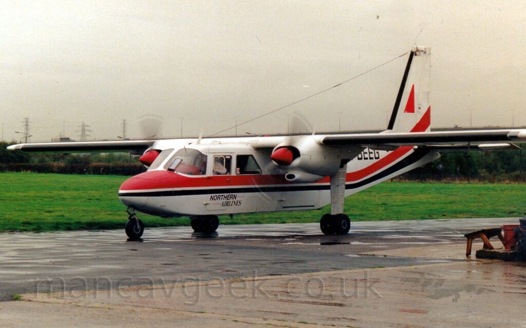 Side view of a small, high-winged, twin propellor-engined airliner parked on grey tarmac apron facing to the left. The plane is largely white, with a thick red and slightly thinner red stripe running along the body below the passenger cabin windows, sweeping up into the lower reaches of the tail from aft of the wing, and small black "Northern Airlines" titles at the bottom of the cockpit doors. The partial black registration "BEEG" can be seen on the upper rear fuselage, while the white tail has a large red triangle in the middle. Grey tarmac apron fills the foreground, with grass in the background leading up to bushes marking the airfield perimeter, under grim grey sky.