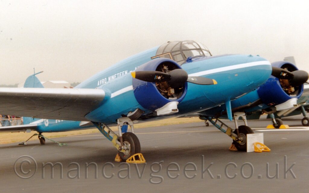 Side view of a blue, twin propellor-engined, World War 2-era transport aircraft parked facing to the right. There is a white stripe running along the body and the wing-mounted engines, with white "Avro Nineteen" titles on the upper fuselage. There are yellow metal chocks in front of and behind the main wheels, with a blue metal support around the tail-wheel.