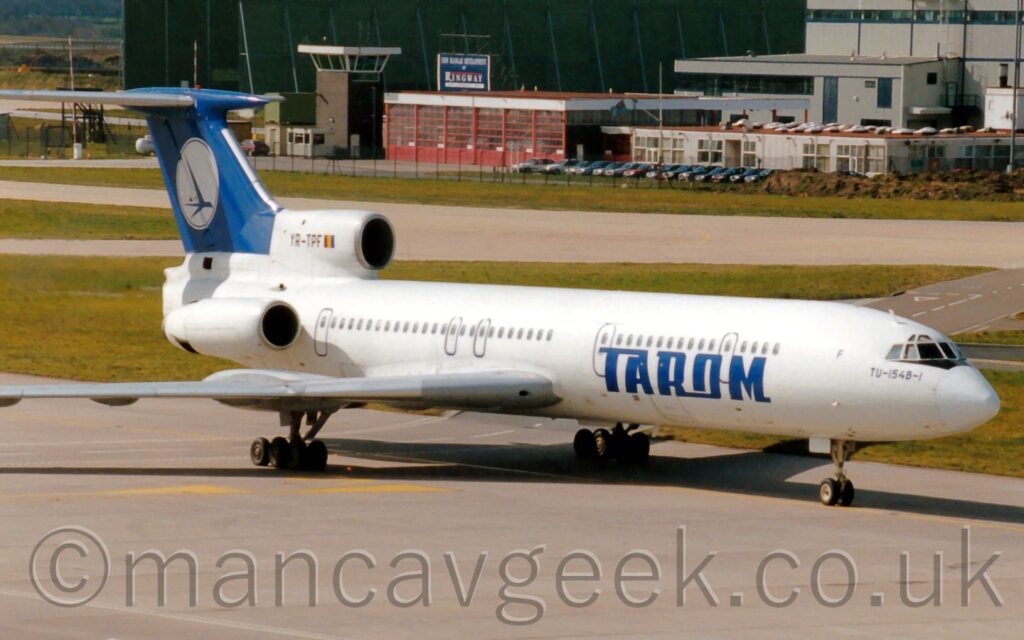 Side view of a white, 3 engined jet airliner with the engines mounted on the rear of the fuselage, taxiing from left to right. There are large blue "Tarom" titles on the lower forward fuselage, and a blue tail with a white circle containing a flying blue bird in the lower right. The registration "YR-TPF" is on the centre engine intake. The plane is taxiing along a large grey taxiway, casting a strong grey shadow forward and to it's left, suggesting strong sunlight coming from low and to the right. Large grass sections separate this taxiway from other taxiways in the background, with grey buildings beyond that. A large, green, metal structure, dominates the rest of the background.