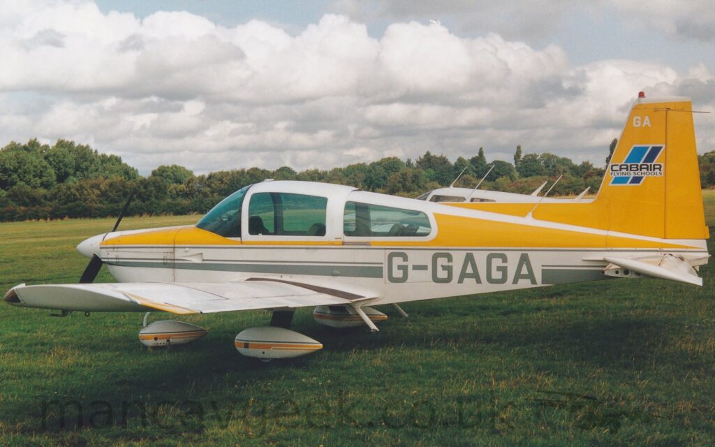 Side view of a single-engined light aircraft parked on grass facing to the left. The plane is largely white, with the top of the engine cowling on the nose, directly in front of the cockpit being yellow, a thi yellow stripe running under the sliding cockpit canopy, and the upper rear fuselage being yellow, outlined in white, and a grey stripe running along the lower fuselage, just above wing level, overlaid with the grey registration "G-GAGA" on the rear fuselage. Yellow and grey stripes also run along the white spats on the landing gear under the nose and wings, and on the tips of the wings. Thae tail, including a large fillet along the top of the fuselage, is also yellow, with a short pair of light and dark blue diagonal bands overlaid with black text "CabAir Flying Schools" in the middle, and the white letters "GA" right at the top, under the red beacon. Green grass fills the foreground, with more grass in the background leading up to trees marking the airfield perimeter. Another light aircraft with a white cockpit roof sits immediately behind this one. A fluffy blanket of white cloud fills the sky, with hints of pale blue occasionally peeking through.