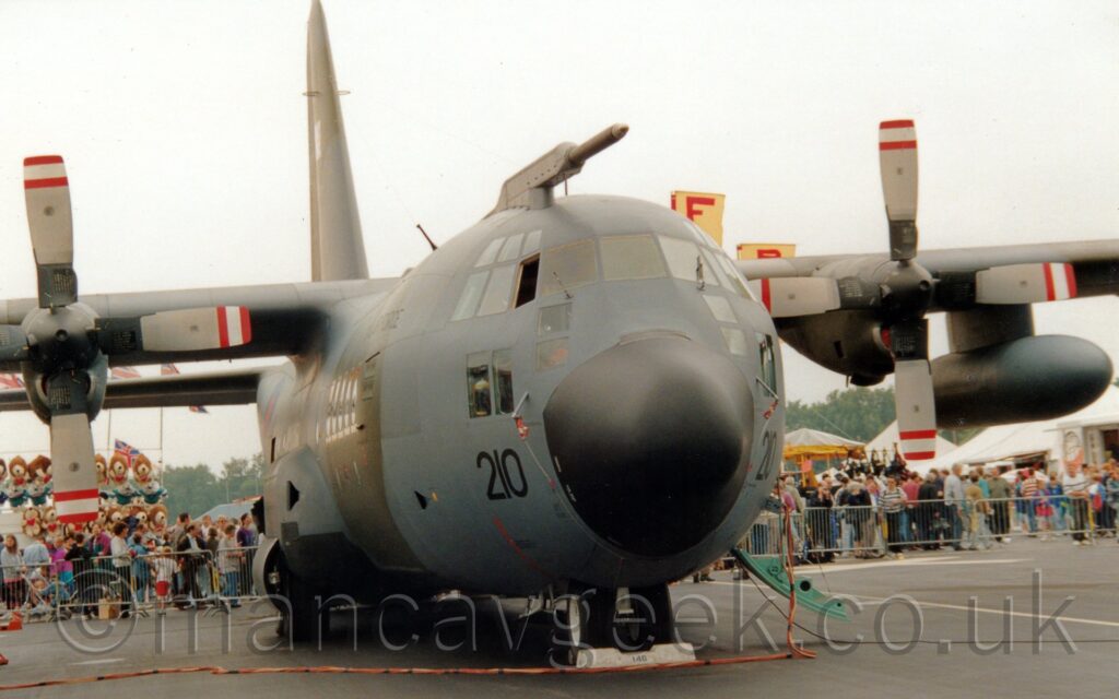 Almost head on view of a high-winged, 4 propellor-engined military transport aircraft parked facing slightly to the right of the camera. The plane has a black radome in the nose, under a heavily glaze cockpit, with a grey and olive green camouflage pattern covering the rest of the plane.T The large black number "210" can be seen on either side of the nose, below the lowest of the cockpit windows, and a large blue and red roundel can be made out on the right rear fuselage. On the far side of the plane, a a door can be seen hanging down from just aft of the cockpit, with stairs formed in the inside. The 4 bladed propellors on the engines are all aligned nicely, in the 4 cardinal positions, with red and white bands on the tips. An orange power cable snakes across the black tarmac the plane is parked on from the left, rising up to plug into an open panel in the nose. In the background, crowds of people can be seen behind temporary metal fences,,,, with various stalls and tents behind them, including one festooned with large brown teddy bears. Flat grey sky fills the rest of the frame.