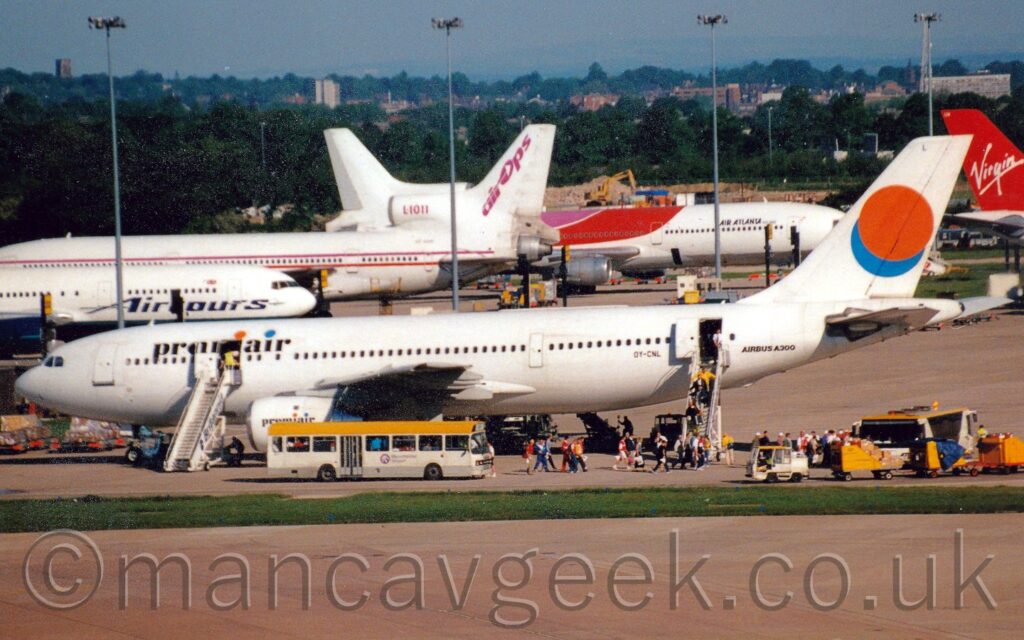 Side view of twin engined jet airliner parked facing to the left. The plane is almost entirely white, with black "PremiAir" titles on the upper forward fuselage, including orange and blue dots over the lower-case letters "i", and the registration "OY-CNL" on the lower rear fuselage in front of the rear passenger door, as well as black text "Airbus A300" after the door. There are also large orange and blue circles on the tail, the orange one partially overlaid on the blue one. The "PremiAir" logo is repeated in a smaller form on the sides of the engine pods under the wings. A white bus with a yellow roof is parked by the engine pods, and another under the planes rear fuselage, with a snake of people walking towards the latter, mostly coming from the airstairs connected to the open rear cabin doors, but also some walking past the other bus. A strip of green grass separates the plane from a large area of concrete apron that fills the foreground, with more apron in the background filles with 4 more large jet airliners parked facing in various directions, all in various shades of white, blue, and red. Tall lighting poles amongst them reach into the sky, with trees and buildings stretch off into the distance, meeting grey sky.