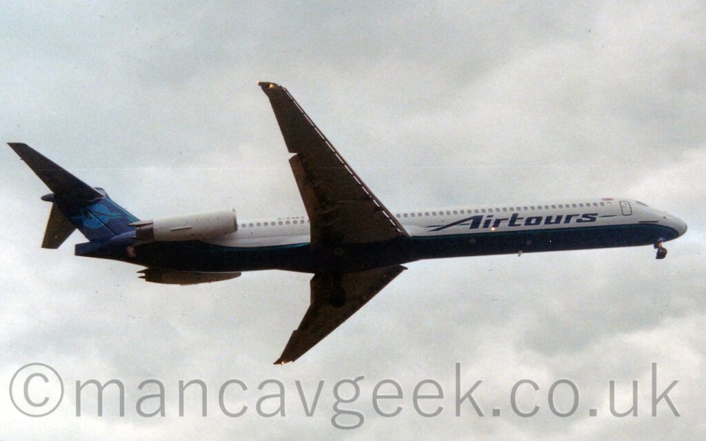 Side view of a twin engined jet airliner with the engines mounted on the sides of the rear fuselage, flying from left to right at a low altitude, with undercarriage lowered and flaps extended from the front and rear of the wings, suggesting it is about to land. The plane is largely white, with a dark blue belly, and a pale green stripe running along the body, both turning upwards behind the engines to fill the tail. There are large, dark blue "Airtours" titles on the lower forward fuselage, and the black registration "G0COES" on the upper rear. A light blue circle outlined in green and containing the dark blue letter "A" is on the dark blue tail. Slightly dramatic white and grey cloudy sky fills the rest of the frame.