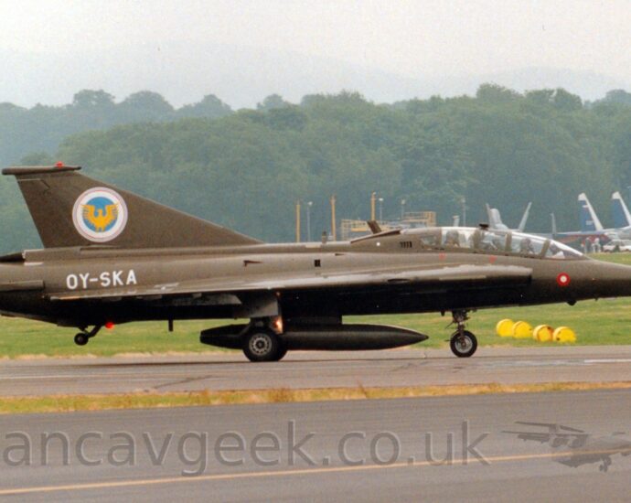 Side view of a military single engined jet fighter taxiing from left to right along a black runway. The plane is in an olive green camouflage, with the white registration "OY-SKA" on the side of the rear fuselage, and a white, blue, and yellow crest on the tail. The plane has a tandem 2 seat cockpit, one pilot sitting behind and above the other. A black taxiway fills the foreground, with grass in the background leading up to trees in the distance, under grey sky.