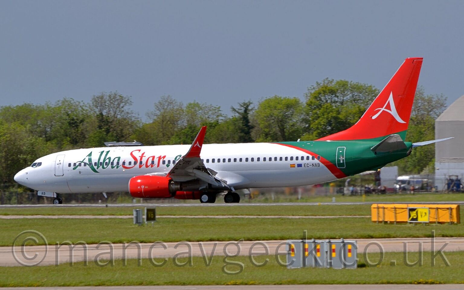 Side view of a twin engined jet airliner moving from right to left along a runway, with flaps drooping from the front and rear of the wings, suggesting it is decelerating after landing. The plane is mostly white, with large green "Alba" and red "Star" titles on the forward fuselage, and the black registration "EC-NAB" on the lower rear fuselage, next to a small Spain and EU flags. The rear fuselage is green with a curved, diagonal red bandtowards the front, while the tail is red with a large, white, stylised letter "A" in the middle, a design replicated on the up-turned wingtips. Red engine pods hang down in front of the wings. Green grass fills the foreground, with grey taxiways passing through from one side to the other. Green trees line the background, under blue-grey skies.