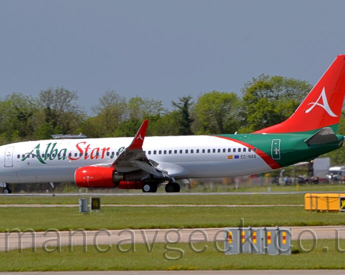 Side view of a twin engined jet airliner moving from right to left along a runway, with flaps drooping from the front and rear of the wings, suggesting it is decelerating after landing. The plane is mostly white, with large green "Alba" and red "Star" titles on the forward fuselage, and the black registration "EC-NAB" on the lower rear fuselage, next to a small Spain and EU flags. The rear fuselage is green with a curved, diagonal red bandtowards the front, while the tail is red with a large, white, stylised letter "A" in the middle, a design replicated on the up-turned wingtips. Red engine pods hang down in front of the wings. Green grass fills the foreground, with grey taxiways passing through from one side to the other. Green trees line the background, under blue-grey skies.