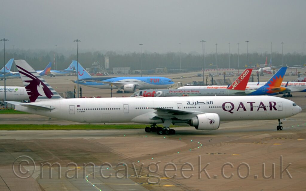 Side view of a twin engined jet airliner taxiing from left to right at a busy airport. The plane is largely white, with mauve, billboard-style "Qatar" titles on the forward fuselage, repeated in a grey Arabic script on the upper forward fuselage, above the wing root, and the black registration "A7-BOF" on the lower rear fuselage, just below the last few cabin windows, and a small Qatari flag just above those windows. The tail is a very light grey, with darker grey stripes overlaid with the head of an animal with horns sticking out of the back of it's head. A smaller, circular version of the tail logo appears on the side of the white engine pods mounted under the wings. Grey concrete apron, marked with yellow lines for planes to follow, some of which are lit up, fills the foreground. More apron space fills the background, with a total of 13 jet airliners visible, parked facing in various directions, in an array of liveries that predominantly feature shades of white, blue, grey, and red. Trees in the distance are being swallowed up by grey mist.