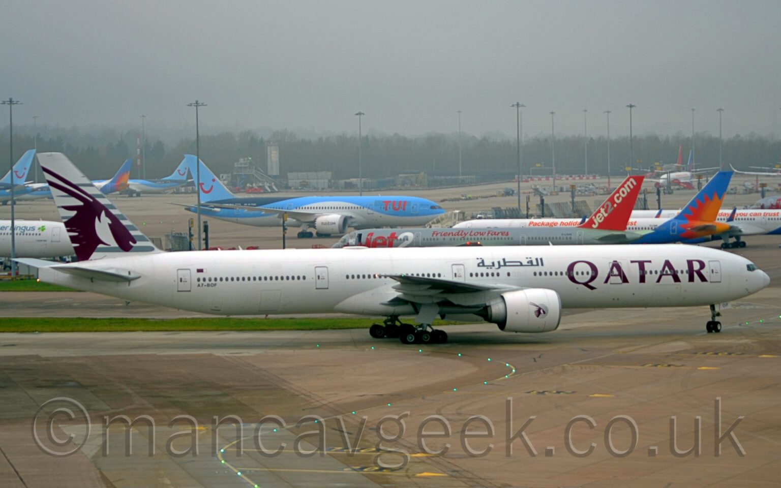 Side view of a twin engined jet airliner taxiing from left to right at a busy airport. The plane is largely white, with mauve, billboard-style "Qatar" titles on the forward fuselage, repeated in a grey Arabic script on the upper forward fuselage, above the wing root, and the black registration "A7-BOF" on the lower rear fuselage, just below the last few cabin windows, and a small Qatari flag just above those windows. The tail is a very light grey, with darker grey stripes overlaid with the head of an animal with horns sticking out of the back of it's head. A smaller, circular version of the tail logo appears on the side of the white engine pods mounted under the wings. Grey concrete apron, marked with yellow lines for planes to follow, some of which are lit up, fills the foreground. More apron space fills the background, with a total of 13 jet airliners visible, parked facing in various directions, in an array of liveries that predominantly feature shades of white, blue, grey, and red. Trees in the distance are being swallowed up by grey mist.