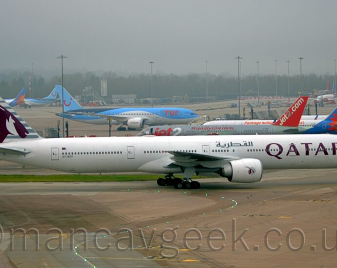 Side view of a twin engined jet airliner taxiing from left to right at a busy airport. The plane is largely white, with mauve, billboard-style "Qatar" titles on the forward fuselage, repeated in a grey Arabic script on the upper forward fuselage, above the wing root, and the black registration "A7-BOF" on the lower rear fuselage, just below the last few cabin windows, and a small Qatari flag just above those windows. The tail is a very light grey, with darker grey stripes overlaid with the head of an animal with horns sticking out of the back of it's head. A smaller, circular version of the tail logo appears on the side of the white engine pods mounted under the wings. Grey concrete apron, marked with yellow lines for planes to follow, some of which are lit up, fills the foreground. More apron space fills the background, with a total of 13 jet airliners visible, parked facing in various directions, in an array of liveries that predominantly feature shades of white, blue, grey, and red. Trees in the distance are being swallowed up by grey mist.