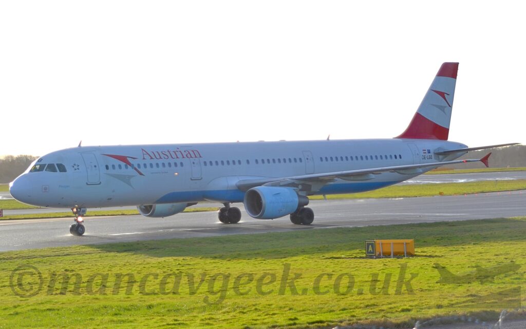 Side view of a twin engined jet airliner taxiing from right to left along a grey taxiway. The plane is mostly white, with red "Austrian" titles behind a stylised flying red bird on the upper forward fuselage a grey shadow of the bird a bit lower down. The black registration "OE-LBB" is on the rear fuselage, just below small Austrian and EU flags. The tail is red at the top and bottom, with a section of a white oval in the middle, with the red bird and grey shadow logo repeated in the middle. The up- and down-turned wingtips are the same red. Green grass fills the foreground, with more grass in the background leading up to trees in the distance, under bright but hazy skies.