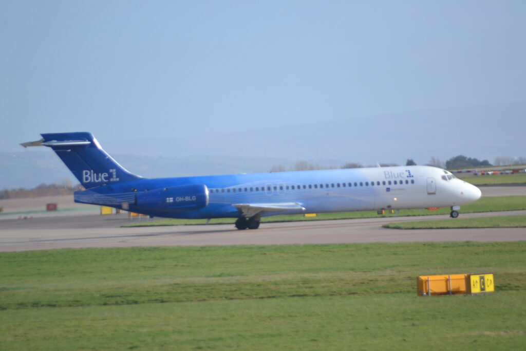 Side view of a twin engined jet airliner with it's engines mounted on the sides of the rear fuselage, taxiing from left to right along a grey taxiway. The plane is white at the front, slowly transitioning through several shades of light blue, to dark blue on the rear fuselage and tail. There are grey "Blue1" titles on the upper forward fuselage, with the number "1" in dark blue, with larger, all grey "Blue1" titles on the base of the tail. The white registration "OH-BLG" is on the side of the blue engine pod, next to a small, white and blue Finnish flag. Green grass fills the foreground, with the yellow backs of some taxiway signs in the middle of the grass. pale blue sky fills the rest of the frame.