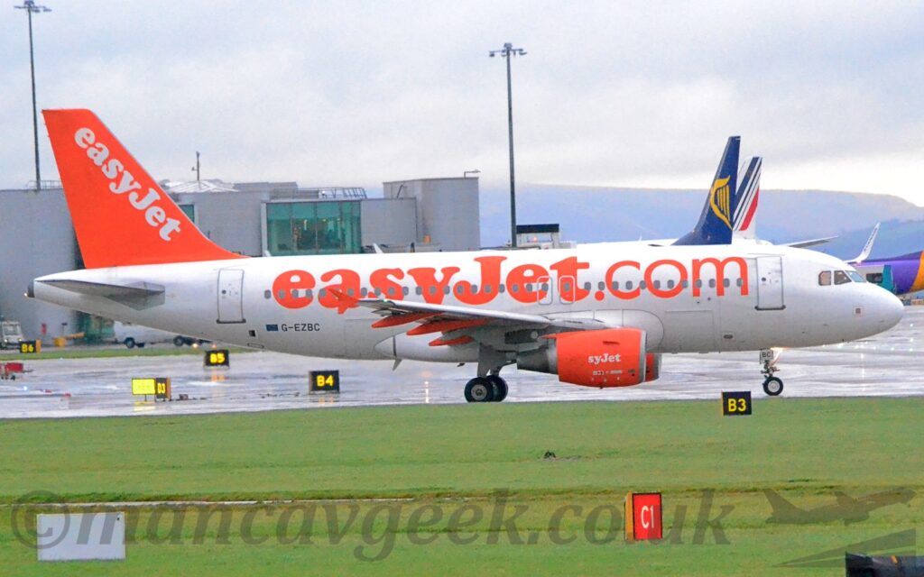 Side view of a twin engined jet airliner taxiing from left to right. The plane is mostly white, with large , billboard-style orange "EasyJet[dot]com" titles on the fuselage, text repeated diagonally in white on the bright orange tail, and on the sides of the engine pods mounted under the wings, although the first 2 letters are missing on the engine. Lush green grass fills the foreground, with a terminal building in the background on the left, and the tails of some other planes on the right. Tree covered hills in the distance meet bright but hazy skies and cloud in the distance.