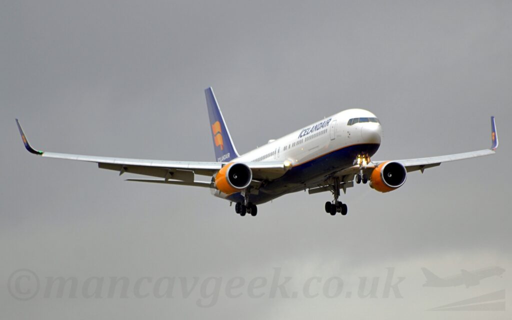 Front view from slightly off to one side of a twin engined jet airliner flying from left to right at a very low altitude, with undercarriage lowered and flaps extended from the front and rear of the wings, suggesting it is about to land. The plane is mostly white, with bright yellow engines, and black "IcelandAir" titles on the upper forward fuselage. The tail is the same dark blue, with a double yellow ribbon curling it's way across the side. The same loge is repeated on both inner and outer faces of the dark blue up-turned wingtips. Flat grey sky fills the rest of the frame.