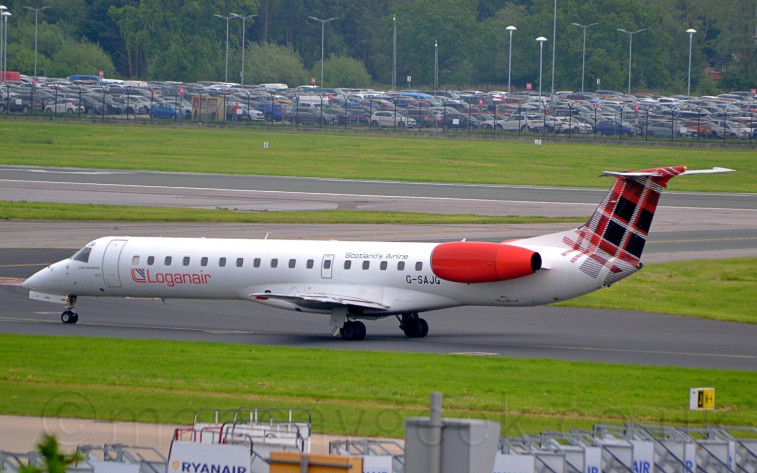 Side view of a white, twin engined jet airliner taxiing from right to left. The plane has it's engines mounted on the sides of the rear fuselage, just below the tail. It is mostly white, with red "LoganAir" titles on the lower rear fuselage, next to a red and black logo which resembles a red, upper-case letter "L" with a black triangle pointing to the upper right. There is a red, black, and white tartan pattern on the tail. Green grass fills most of the foreground, with a collection of mobile airstairs right at the bottom of the frame. Taxiways and a runway lined with green grass, in the background lead up to a nearly full car park surrounded by a black metal mesh fence, with tall lighting poles scattered amongst the cars, with tall trees beyond.