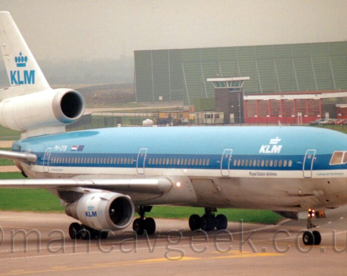 Side view of a 3 engined jet airliner taxiing from left to right along a grey concrete taxiway. The plane is pale blue on the top, with a darker blue stripe covering the passenger cabin windows, and a thinner white stripe, with the lower body in a rather battered bare metal finish. There are white letters "KLM" under a stylised white crown on the upper forward fuselage, above blue text "Royal Dutch Airlines" on the white stripe below. The white registration "PH-BTD" is on the upper rear fuselage, next to small Dutch and EU flags. The "KLM" crown logo is repeated on the sides of the white engine pods under the wings, this time in blue, as well as in a larger blue form on the white tail, above the white centre engine pod. Grey concrete apron fills the foreground, with more taxiways criss-crossing a grassed area in the background, in front of a red, heavily glazed building with a squat brown tower with a heavily glazed top floor, which itself sits in front of a large, green, box-like structure. Dreary, cloudy grey sky fills the rest of the frame.