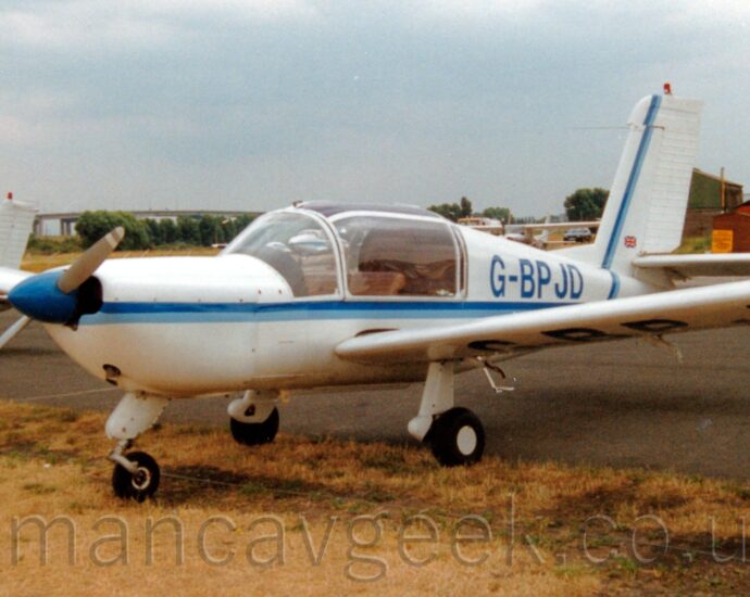 Side view of a single-engined light aircraft parked facing to the left on yellowing grass, right on the edge of a black tarmac taxiway. The plane is mostly white, with a 2-tone blue stripe running along the body before sweeping up the front of the tail. The registration "G-BPJD" is on the upper rear fuselage, as well as under the left wing. Another couple of planes are parked around it, as well as a couple of wooden buildings on the right. A long bridge reaches over the horizon on the left, with blue-grey sky filling the rest of the frame.