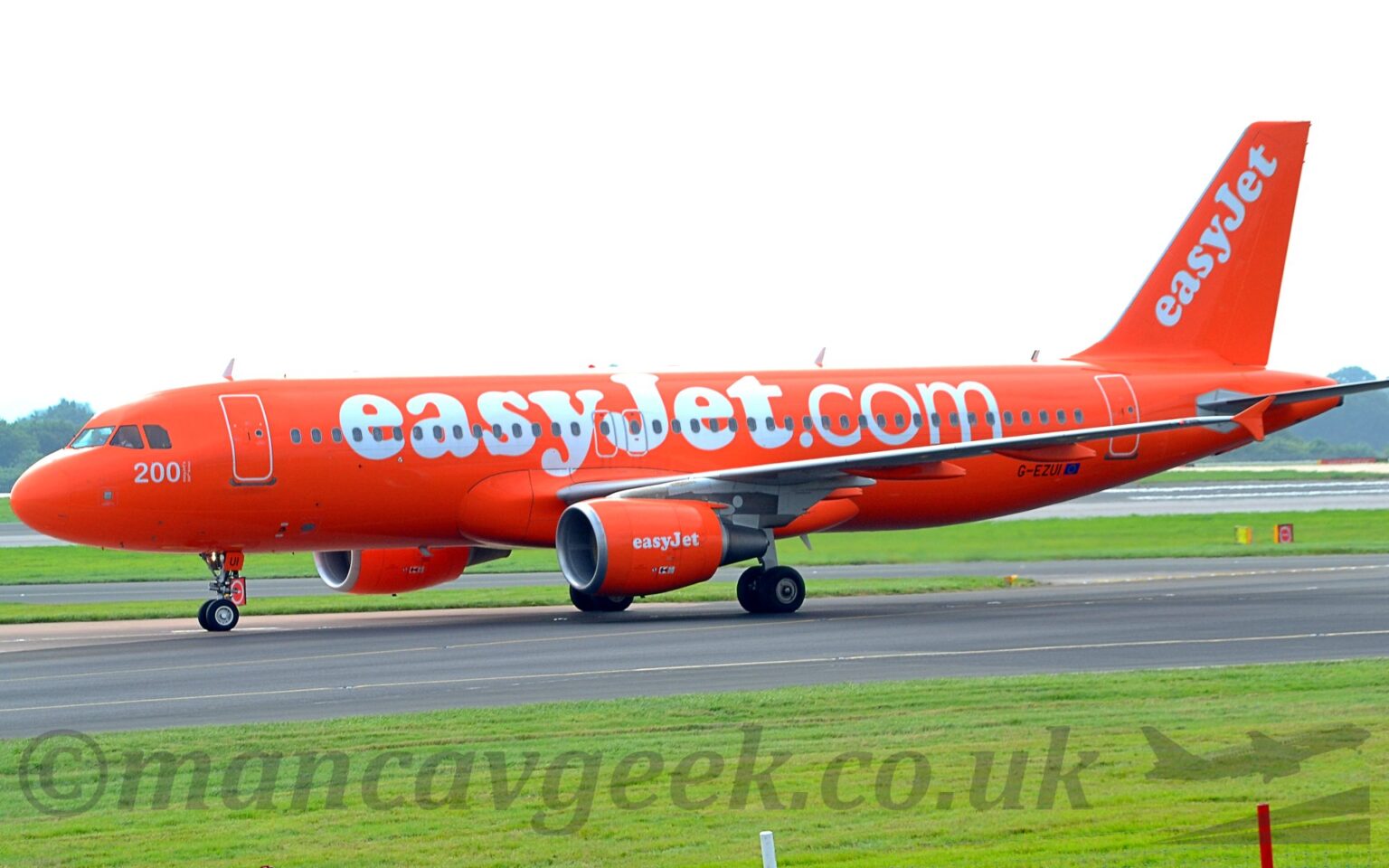Side view of an extremely orange, twin engined jet airliner taxiing from right to left along a black taxiway. The plane is almost entirely orange, with billboard-style white "easyjet.com" titles on the side of the fuselage, the white number "200" under the cockpit windows, and diagonal white "easyJet" titles on the tail. Even the wingtip fences are painted orange, while the black registration "G-EZUI" is the only break in the colour scheme. Lush green grass fills the foreground, with more visible under the plane in the background, and trees on the skyline in the distance. Flat white sky fills the top of the frame.