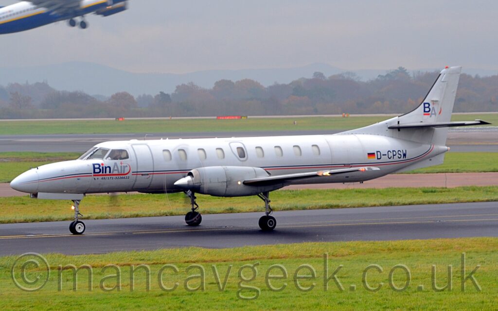 Side view of a long and thin, twin propellor-engined airliner taxiing from right to left along a black tarmac taxiway. The plane is largely white, with a thin blue and red stripe running along the body, overlaid with blue "BinAir" titles under the cockpit windows, and the black registration "D-CPSW" next to a small German flagon the side of the rear fuselage. The white tail has the blue letters "BA" underlined in red. Lush green grass fills the foreground, with more grass in the background leading up to a black runway , and more grass leading up to trees in the distance, under grim grey sky. The centre fuselage and wingbox of a white, yellow, and blue jet airliner flying through the top left corner as it takes off.