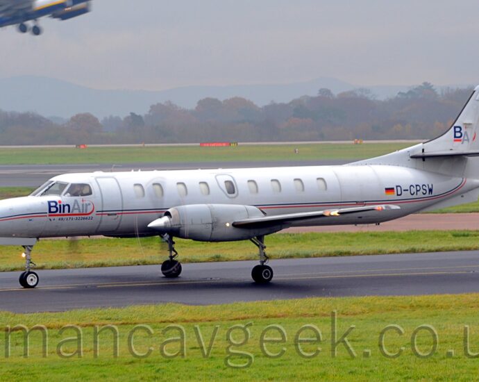 Side view of a long and thin, twin propellor-engined airliner taxiing from right to left along a black tarmac taxiway. The plane is largely white, with a thin blue and red stripe running along the body, overlaid with blue "BinAir" titles under the cockpit windows, and the black registration "D-CPSW" next to a small German flagon the side of the rear fuselage. The white tail has the blue letters "BA" underlined in red. Lush green grass fills the foreground, with more grass in the background leading up to a black runway , and more grass leading up to trees in the distance, under grim grey sky. The centre fuselage and wingbox of a white, yellow, and blue jet airliner flying through the top left corner as it takes off.