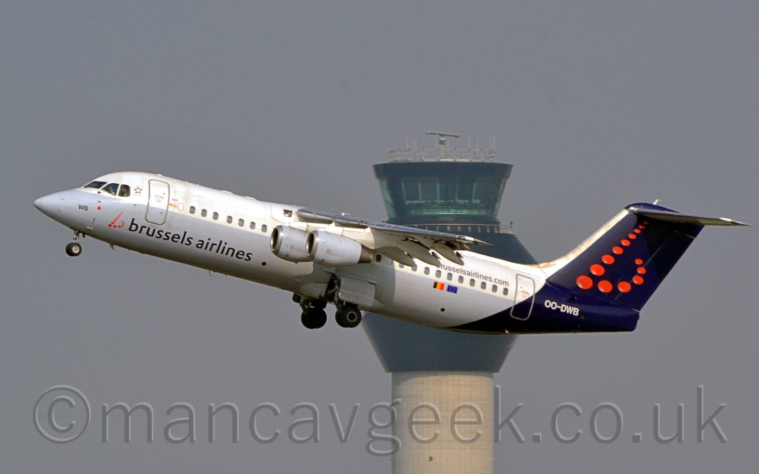 Side view of a high-winged, 4 engined jet airliner flying from right to left at a low altitude, with undercarriage extended, flaps extended from the rear of the wings, and the nose raised sharply, suggesting it has just taken off. The plane is white on top of the body and grey on the lower half, with dark blue, almost purple "Brussels Airlines" titles just aft of a stylised red lower-case letter "b" made upe of red dots. The tail and rear fuselage is the same dark blue, with a larger version of the red dot "b" logo on the tail, above the white registration "OO-DWB" on the fuselage. The web address "BrusselsAirlinesDotCom" is on the upper rear fuselage, above the rear cabin windows, with small Belgian and EU flags below those windows. Grey engine pods hang down below the wings. A tall grey tower with a black top reaches into the sky in the middle of the background, with grey-blue sky filling the rest of the frame.