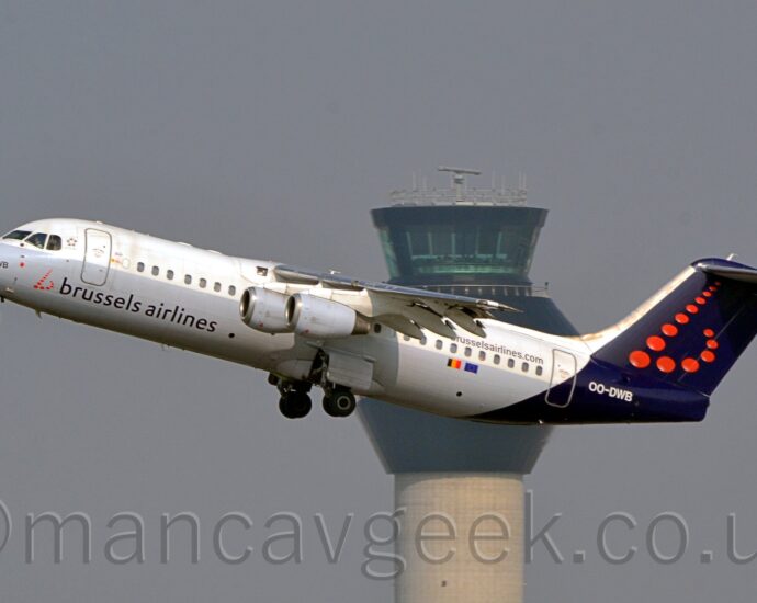 Side view of a high-winged, 4 engined jet airliner flying from right to left at a low altitude, with undercarriage extended, flaps extended from the rear of the wings, and the nose raised sharply, suggesting it has just taken off. The plane is white on top of the body and grey on the lower half, with dark blue, almost purple "Brussels Airlines" titles just aft of a stylised red lower-case letter "b" made upe of red dots. The tail and rear fuselage is the same dark blue, with a larger version of the red dot "b" logo on the tail, above the white registration "OO-DWB" on the fuselage. The web address "BrusselsAirlinesDotCom" is on the upper rear fuselage, above the rear cabin windows, with small Belgian and EU flags below those windows. Grey engine pods hang down below the wings. A tall grey tower with a black top reaches into the sky in the middle of the background, with grey-blue sky filling the rest of the frame.