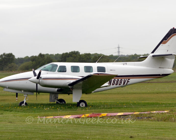 Side view of a twin propellor-engined parked facing to the left on grass. The plane is largely white, with a black and brown stripe running along the body and engine cowlings, as well as along the top of the tail. The black registration "N889VF" is on the lower rear fuselage with a brown drop shadow. Green grass fills the foreground, with more grass leading up to trees in the distance under flat grey sky, with a tall electricity pylon reaching into the sky, with wires stretching across the width of the frame.
