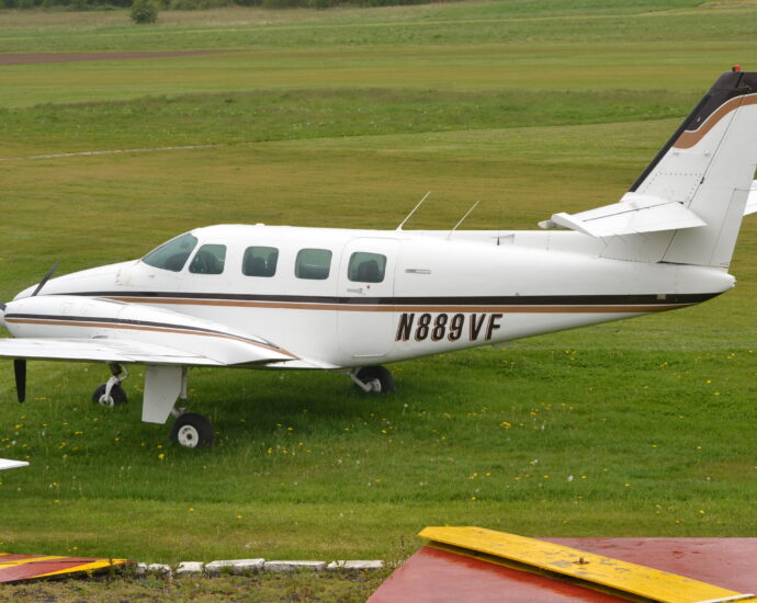 Slightly elevated side view of a twin propellor-engined BizProp parked on grass facing to the left. The plane is largely white, with a black and brown stripe running along the body, as well as along the sides of the engine cowling on the wings. The black registration "N889VF" is under the stripe on the lower rear fuselage, with a brown drop shadow. Green grass fills most of the frame, mostly short cropped, with some slightly longer patches, and with some trees right at the back.