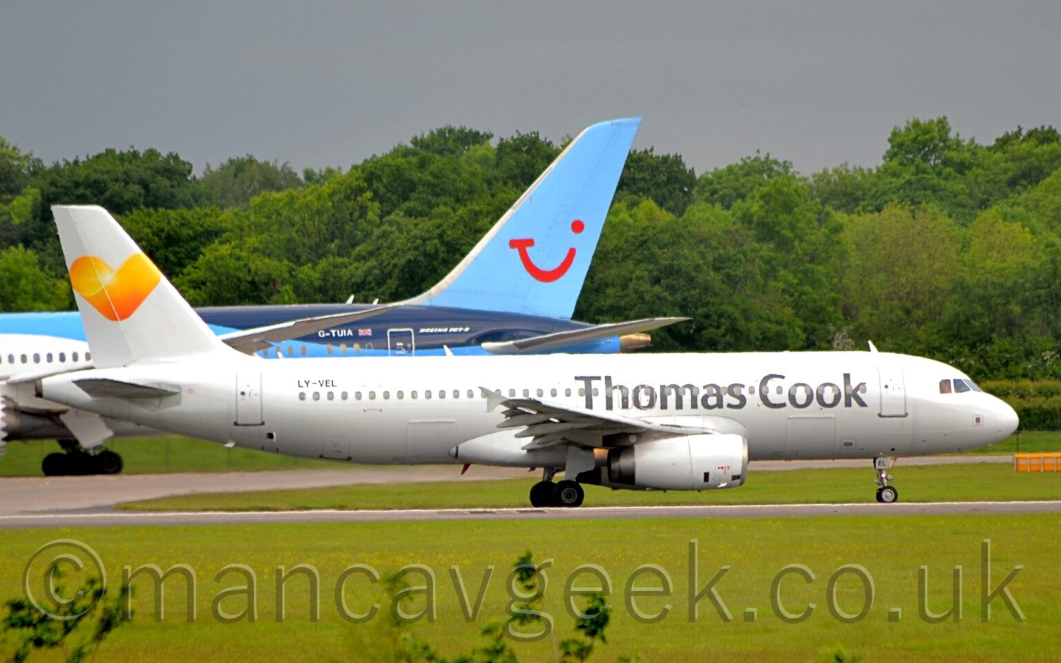 Side view of a white, twin engined jet airliner taxiing from left to right. The plane has large, grey, billboard-style "Thomas Cook" titles on the forward fuselage, and a yellow love heart logo on the tail. Lush green grass fills the foreground, while in the background, the dark blue, light blue, and white rear fuselage and tail of another jet airliner can be seen on the left. Green trees stretch across the frame in the distance, under steely grey sky.