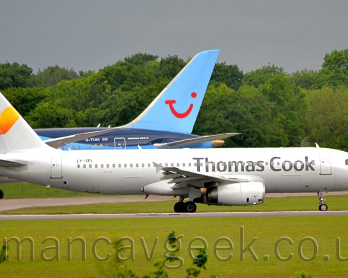 Side view of a white, twin engined jet airliner taxiing from left to right. The plane has large, grey, billboard-style "Thomas Cook" titles on the forward fuselage, and a yellow love heart logo on the tail. Lush green grass fills the foreground, while in the background, the dark blue, light blue, and white rear fuselage and tail of another jet airliner can be seen on the left. Green trees stretch across the frame in the distance, under steely grey sky.
