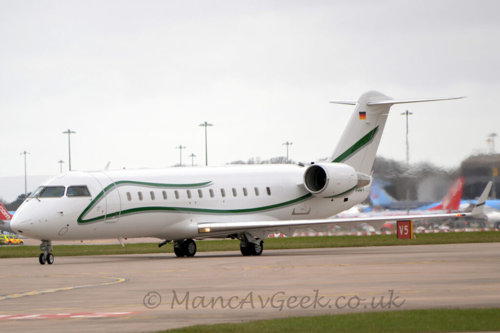 Side view of a twin engined BizJet with the engines mounted on the sides of the rear fuselage, taxiing from right to left along a grey taxiway. The plane is largely white, with a green stripe starting in the middle of the upper fuselage, running forward along the top of the body, down under the cockpit windows, then back along the lower body, and up into the tail, and the green registration "D-AJOY" on the tail-cone, partially obscured by one of the engines on the rear fuselage. A grey taxiway fills the foreground, with a patch of green grass in the bottom right corner, and more grass in the background, leading up to several jet airliners partially obscured by jelly pouring out of the rear of the planes engines. Flat grey sky fills the rest of the frame.