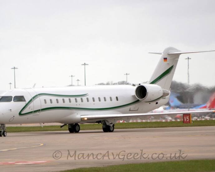 Side view of a twin engined BizJet with the engines mounted on the sides of the rear fuselage, taxiing from right to left along a grey taxiway. The plane is largely white, with a green stripe starting in the middle of the upper fuselage, running forward along the top of the body, down under the cockpit windows, then back along the lower body, and up into the tail, and the green registration "D-AJOY" on the tail-cone, partially obscured by one of the engines on the rear fuselage. A grey taxiway fills the foreground, with a patch of green grass in the bottom right corner, and more grass in the background, leading up to several jet airliners partially obscured by jelly pouring out of the rear of the planes engines. Flat grey sky fills the rest of the frame.